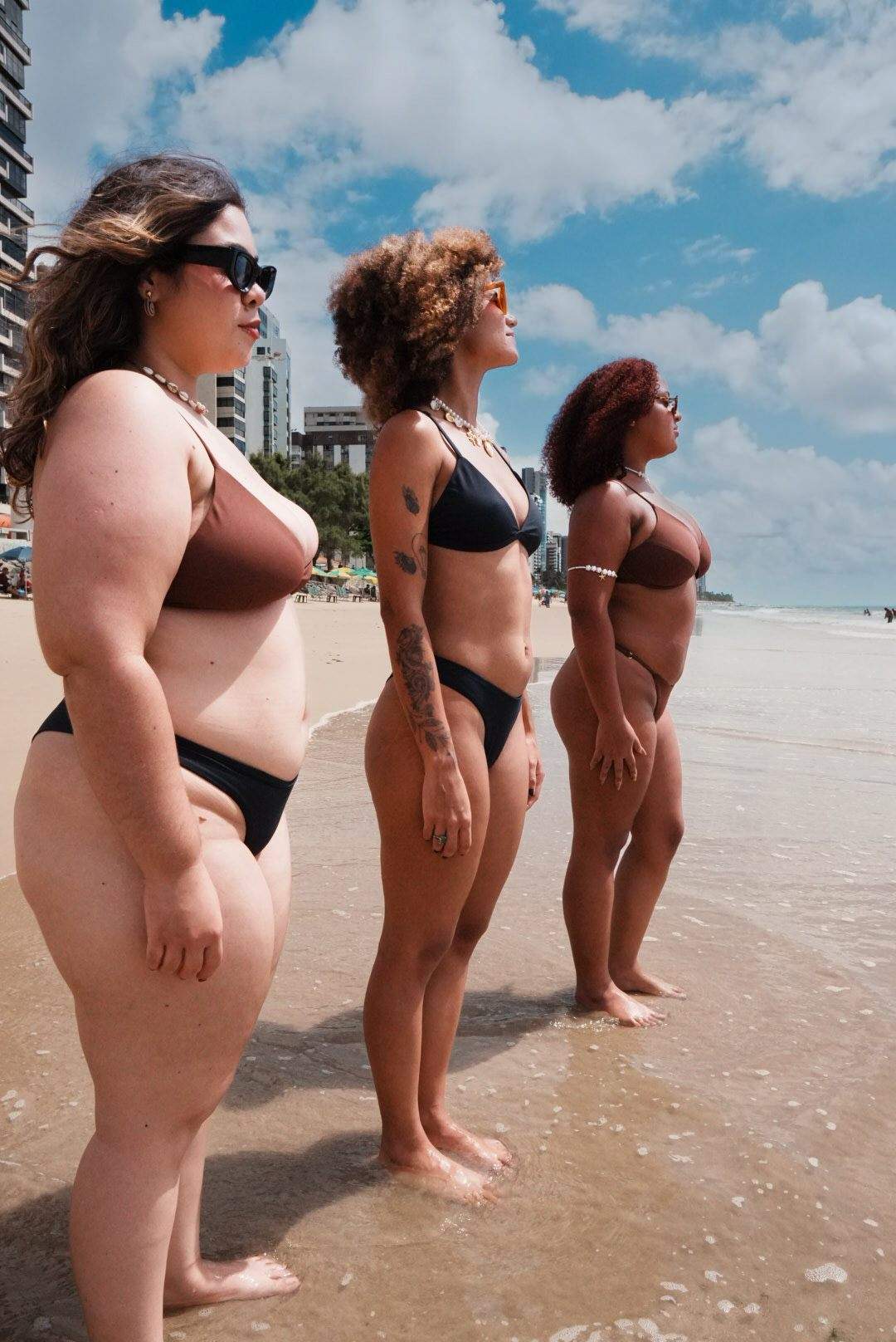Três mulheres de biquíni, lado a lado na beira do mar